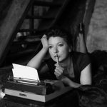 A reflective woman poses elegantly with a typewriter in a black and white setting, evoking nostalgia.