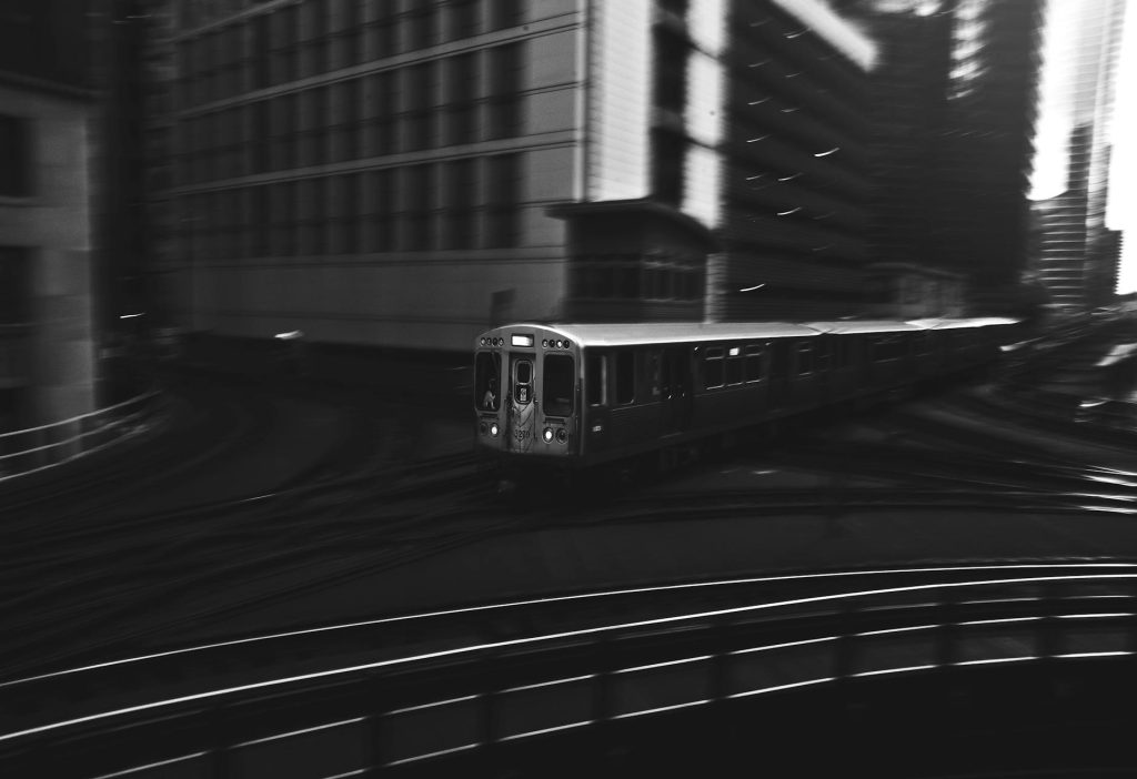 Dynamic black and white photo of a train navigating through a bustling cityscape at night.