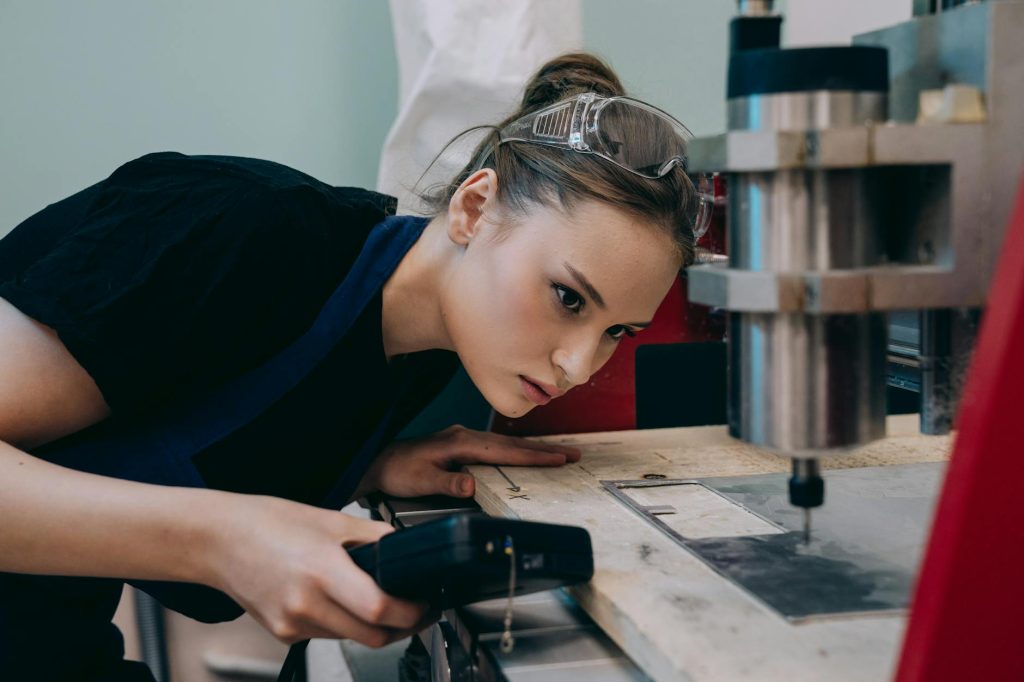 Woman closely inspecting machinery in a workshop wearing protective goggles.