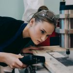 Woman closely inspecting machinery in a workshop wearing protective goggles.