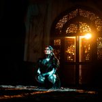 A woman in black attire kneels in dramatic lighting with ornate door backdrop.