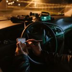 A person in a car at night, writing notes, with binoculars and camera on dashboard.