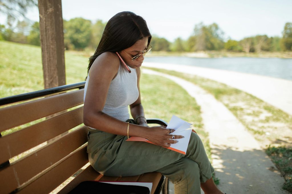 A woman is sitting on a bench outdoors, reading and reviewing documents by a scenic path.