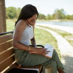 A woman is sitting on a bench outdoors, reading and reviewing documents by a scenic path.