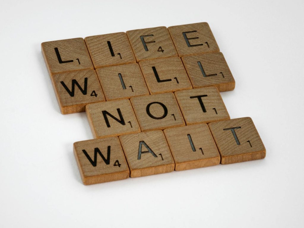 Wooden Scrabble tiles spelling 'Life Will Not Wait' on a white background.