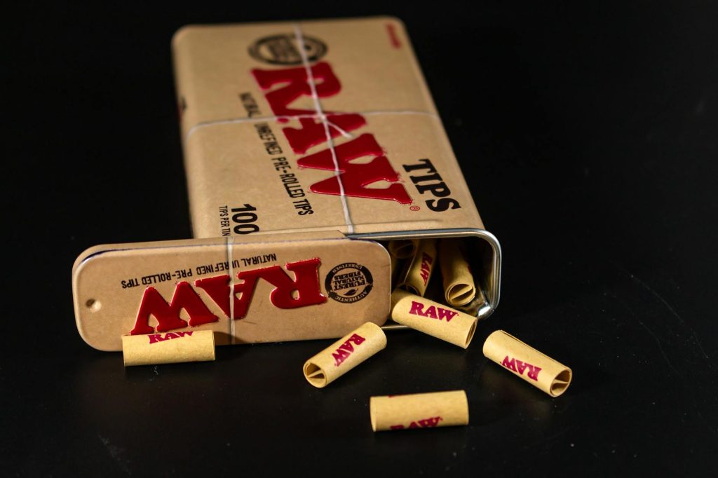 Close-up of RAW pre-rolled tips spilling from a metal tin on a black surface.