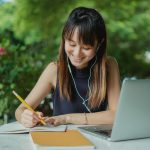 Positive young Asian female student with earphones writing in copybook while doing homework at table with laptop in street cafeteria
