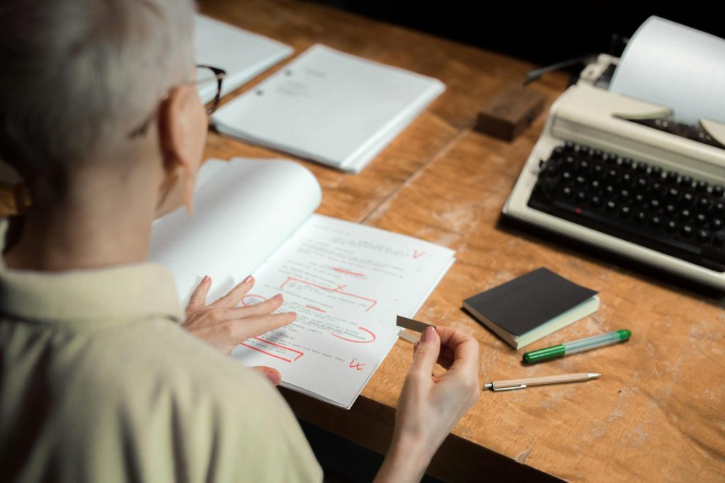 An adult woman reviewing a script with red pen marks at a wooden desk with a typewriter.