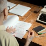 An adult woman reviewing a script with red pen marks at a wooden desk with a typewriter.