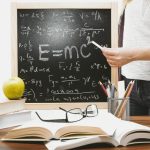 Woman writing physics equations on a blackboard with books and an apple on the desk.