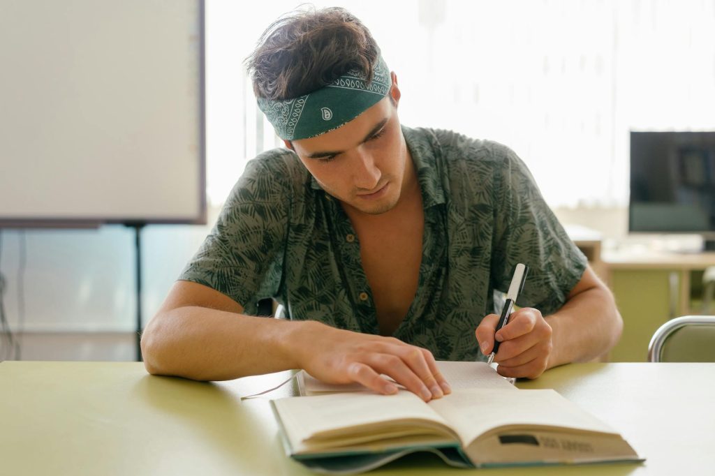 Young man intensely studying with a notebook at a desk indoors, focusing on research and writing.
