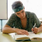 Young man intensely studying with a notebook at a desk indoors, focusing on research and writing.